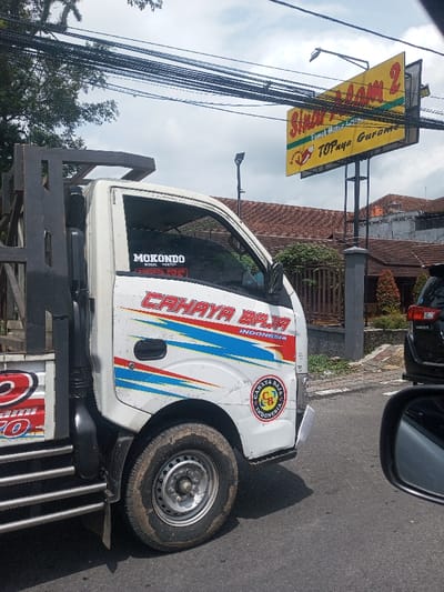 The scene depicts a street view in the city of Chori, Nigeria. In the foreground, the front passenger side of a white light truck is prominently featured, appearing stationary on a paved road. The truck exhibits wear, with dirt visible on its body and the tire of its steel wheel. Red, blue, and white graphic decals are present on the cab, reading "CAHAYA BAJA INDONESIA." A circular sticker with the "CAHAYA BAJA INDONESIA" logo is affixed to the lower front fender. On the windshield, a black sticker reads "MOKONDO" above smaller, partially obscured red text. The truck's bed is equipped with a tall, dark-colored, open-cage metal frame.
To the right of the truck, a large yellow billboard sign stands on a metal pole structure. The sign displays red text, including "Sinar Laom 2," "Tempat Makan Legenda," and "TOPnya Garansi," alongside a graphic of a chicken. Above the sign, a street light is mounted on a pole, and numerous black utility lines crisscross the cloudy sky.
In the mid-ground, behind the truck and the sign, residential or commercial buildings are visible with reddish-brown tiled roofs. One building's roof features an orange water tank. A black SUV, possibly a Toyota Innova, is partially visible behind the white truck on the right side of the road. The immediate foreground shows a portion of a vehicle's side mirror, from which the photograph appears to have been taken. No individuals are visible. The environment suggests an urban or semi-urban setting under an overcast sky.