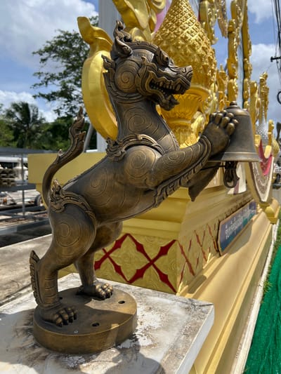 A medium shot captures a golden statue of a mythical creature, possibly a lion or griffin, holding a bell in Pa Khlok, Thailand. The statue is positioned outdoors against a backdrop of bright blue sky with scattered white clouds and lush green trees. The creature is rendered in a bronze-like material with intricate swirling patterns and ornate detailing on its body, limbs, and tail. Its mouth is open in a fierce expression, revealing sharp teeth. The statue stands on a circular base, which is placed on a weathered concrete pedestal. Behind the statue, a vibrant yellow structure with red and gold decorative elements suggests it is part of a temple or shrine. A blue sign with Thai script is visible on the yellow structure. The overall scene is well-lit, indicating a sunny day.
