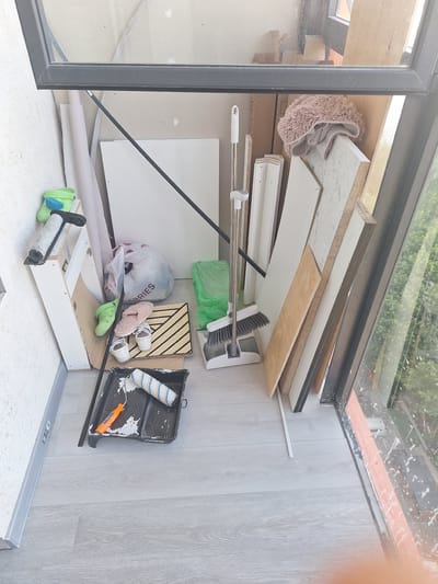 The image shows a cluttered corner, possibly on a balcony or in a storage area, with various construction and household items. The floor is light gray wood-look laminate. Against a textured white wall, a white wooden shelf unit holds two pairs of green clogs and a roller for painting with a black handle and a white roller with blue and yellow stripes sits in a black paint tray with white paint splatters on the floor around it. To the right of the shelf unit are several vertical white boards and a light brown wooden board leaning against each other. A white mop with a broom attachment stands upright. A crumpled pink fluffy blanket is draped over a piece of wood. A clear plastic bag with the word "RIES" visible on it is on the floor. A green plastic bag is also on the floor. Two pairs of slippers, one pink fluffy and one white with straps, are next to a square mat with diagonal stripes. The scene is viewed through a dark metal frame, possibly of a window or doorway, with reflections visible on the glass. The overall impression is one of disarray, with items seemingly stored temporarily or in the process of being used. No specific details of Balashikha, Russia, are discernible from the image.