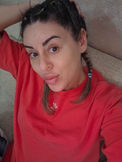 A medium shot captures a woman in her late 30s or early 40s from a high angle, lying down on a beige sofa. Her head is tilted back, resting on her left arm, which is bent at the elbow and positioned behind her head. Her right arm is not visible. She has dark brown hair styled in two braids, one on each side, with a few strands loose around her face. She is wearing a bright red sweatshirt with a white Adidas logo on the left chest. Her facial expression is playful, with her lips pursed and her eyes looking directly at the camera. The background consists of a textured wall and the sofa she is lying on. The scene appears to be indoors, likely in a residential setting. There are no other individuals or objects of significance in the frame. The lighting is soft and even, suggesting natural light or diffused indoor lighting.