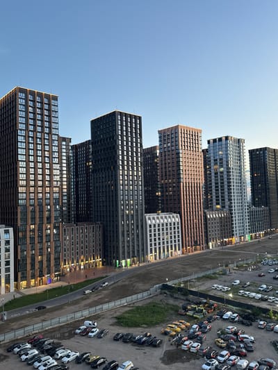 The image shows a contemporary urban landscape with numerous high-rise residential buildings under a clear blue sky. The buildings vary in architectural style and facade materials, with some featuring brickwork and others glass and metal. Many windows reflect the sunset, casting warm tones onto the structures. In the foreground, a large, partially developed area is visible, with a road running alongside the buildings. This road is lined with streetlights and appears to have some pedestrian activity. Beyond the road, a vast parking lot is filled with a variety of vehicles, including cars, vans, and construction equipment such as excavators and a road roller. The scene suggests a newly developed or developing urban area, possibly on the outskirts of a city. The location is identified as Bobrovo, Russia.