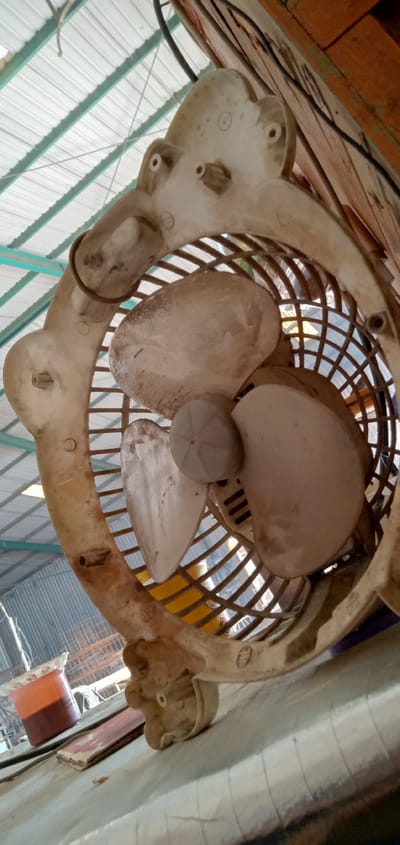 The image depicts a dirty, white plastic electric fan with three blades, encased in a round cage, in Banjaran, Indonesia. The fan appears to be old and in need of cleaning, as evidenced by the dust and grime accumulated on its surfaces. The fan is positioned indoors, possibly in a workspace or storage area, with a glimpse of a corrugated metal roof and surrounding objects suggesting a cluttered environment. A container of liquid is on the surface.