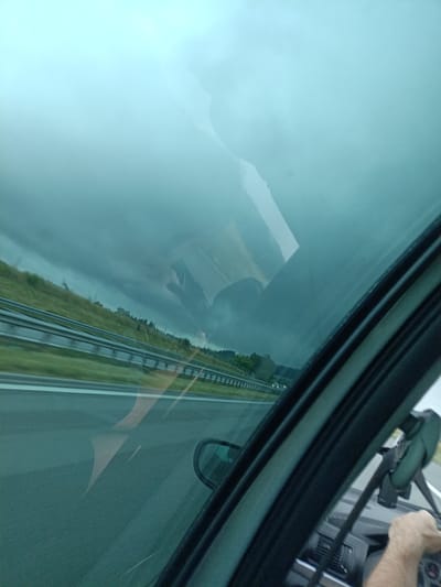 The image captures a view from inside a car, looking through the windshield. The scene depicts a highway with a guardrail and a grassy embankment to the left, suggesting a rural or suburban setting. The sky is overcast and appears to be a stormy gray, hinting at impending rain or a recent storm. The overall lighting is dim, characteristic of a cloudy or twilight day.
Reflected in the windshield are images of the car's interior and its occupants. We can see a hand on the steering wheel, indicating someone is driving. There are also reflections of what appear to be heads, possibly of passengers, though their features are indistinct due to the reflections and the dim lighting. A dashcam is mounted on the dashboard, partially visible.
The road ahead shows lane markings and other vehicles in the distance, suggesting active traffic. The greenery on the embankment appears lush, indicating it's likely springtime or summer. The strong blue tint of the windshield modifies the colors of the scene, giving it a slightly surreal or moody atmosphere. The overall impression is one of traveling through a potentially dramatic weather event.
