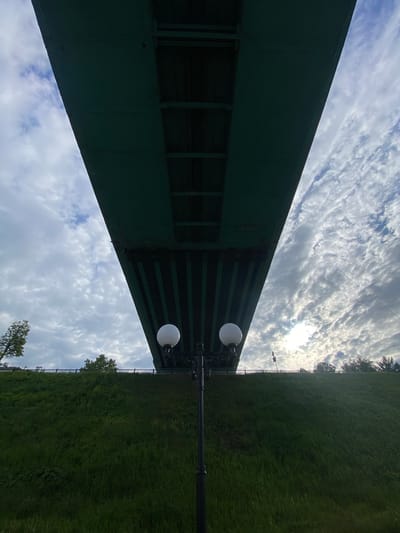 The image displays an outdoor scene featuring a large, dark green bridge or overpass structure positioned across the upper left and central portions of the frame. This structure has visible support beams and horizontal elements underneath.

To the right of the green structure, a black lamppost with an ornate top stands, fitted with two spherical white light fixtures. Beyond the lamppost and spanning the lower right of the image, a lush green grassy hillside or embankment extends, dotted with trees along its distant horizon line.

The sky, visible above and to the right of the bridge and behind the lamppost and trees, is partly cloudy with white, scattered clouds. Bright sunlight filters through the clouds, creating a radiant area in the upper right. The scene is set during the daytime under bright, indirect sunlight. No people or visible text are present. The location appears to be a public outdoor area, possibly a park or riverside setting adjacent to an elevated roadway or railway.