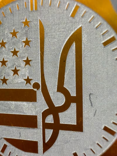 This is a macro photograph of a medal or coin, focusing on its intricate design. The medal is silver with gold accents and features a series of markings along its outer edge, resembling notches or indicators. The central design is a stylized, abstract symbol in gold. Below this symbol, a pattern of gold stars is visible, reminiscent of the American flag, arranged in rows. The surface of the medal exhibits a brushed texture, and there are some faint scuffs or marks on its surface. The background is out of focus, appearing as a warm, golden hue, possibly indicating the medal is resting on a similar colored surface or that light is reflecting from such a surface. The image captures the detail of the engraving and the contrast between the silver and gold elements. There is no visible text. The context of Grapevine, United States of America, suggests this might be a commemorative item related to an event or organization in that location.