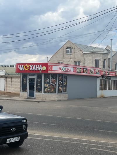 A storefront with red signage reading "ЧАХАНА" in Cyrillic, featuring an image of a ladle and a stylized pepper. Below the sign, a display window showcases images of various dishes. The building has a grey garage door and a white brick facade. A black car with a license plate reading "K 005 BY 05" is parked in the foreground. The sky is overcast with visible power lines. The scene appears to be in a commercial area on the outskirts of a city, possibly in Russia, given the Cyrillic signage and license plate format.