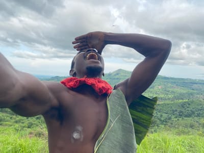 The image shows a close-up, upside-down view of a person in an outdoor setting. The subject is a dark-skinned individual with a mustache and open mouth, revealing teeth. They are adorned with a red fabric around their neck and a large green leaf draped over their shoulder and chest, which appears to be a banana leaf. White markings are visible on the subject's shoulder and chest. The background is a blurred landscape with lush green grass and hills, suggesting a rural or natural environment. The sky is cloudy, indicating overcast weather. The image orientation is inverted, with the sky appearing at the bottom and the landscape at the top. The overall impression is of a candid shot capturing a moment of expression from the subject. The location is identified as Asaga Amangwu, Nigeria. The time of day is not explicitly indicated but the lighting suggests daytime. No visible text is present in the image.