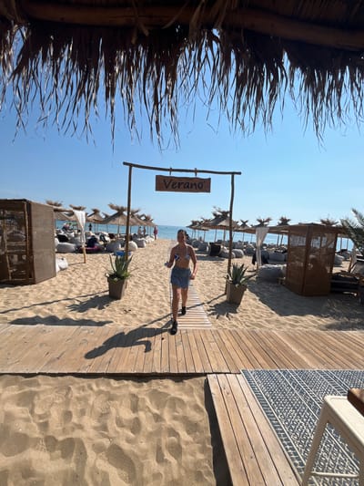 A young woman with dark hair, wearing a blue and white patterned bikini top and a denim skirt, walks on a wooden pathway on a sandy beach. She is in the foreground, facing slightly away from the camera and looking towards the right. Behind her, a wooden archway with a sign that reads "Verano" is visible. The beach is lined with thatched umbrellas and beanbag chairs, suggesting a resort or lounge area. In the background, the sea and a clear blue sky are visible. The scene appears to be in Dyuni, Bulgaria, as indicated by the context. The lighting suggests it is daytime.