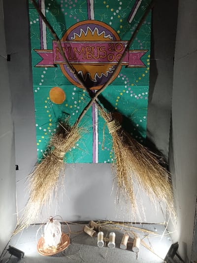 The image displays a Harry Potter-themed exhibit located in El Port De Sagunt, Spain, set against plain gray walls and illuminated by two floor-level spotlights. Centered vertically is a large rectangular banner, appearing to be made of corrugated cardboard, painted teal green with white, purple, and orange accents. The banner features a central orange circle with white "sunburst" patterns, overlayed by a pink banner bearing the text "NIMBUS 2000" in an ornate font. The green background is speckled with small white, yellow, and purple dots, and a single orange circle is present on the lower left of the banner.
Two rustic brooms, constructed from natural branches as handles and bundles of dried straw or grass tied with twine, are prominently displayed. They cross in an 'X' formation over the banner, with their straw ends extending downwards.
On the gray floor in the foreground, an arrangement of objects is present. To the left, a white, stylized owl figure, possibly made of paper or ceramic, is contained within a copper-colored wire cage, which rests on a matching copper-colored circular tray. To the right of this, several cardboard tubes are arranged; some are plain cardboard, while others feature silver-colored end caps. Scattered dry straw or hay is dispersed across the floor, particularly beneath the brooms and around the foreground objects. The two visible spotlights positioned on the floor cast direct light onto the display from the left and right, creating shadows. No individuals are present in the scene.