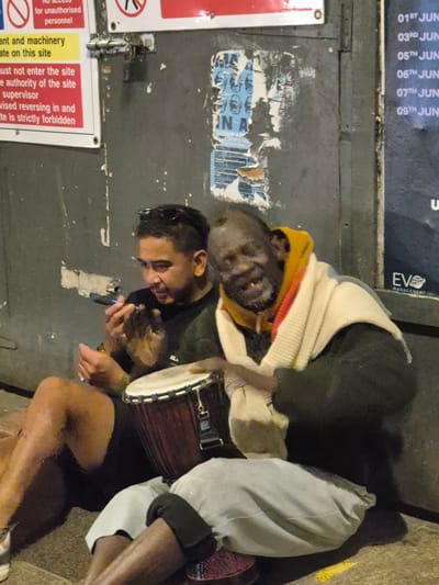 The image depicts two men situated next to a corrugated metal door in Cardiff, United Kingdom. One man is playing a djembe drum. The other man appears to be holding a phone. A sign affixed to the door reads "No access for unauthorised personnel". Additional text is visible on the sign, cautioning against entering the site without supervision and forbidding reversing. A partial view of a poster indicating dates in June is visible at the top of the image.