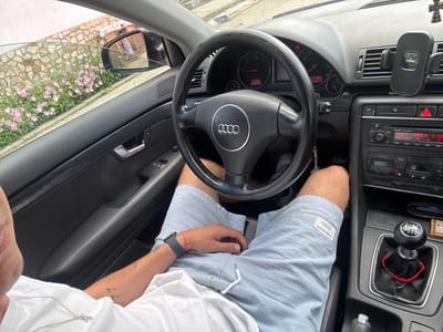 A first-person perspective photo depicts a person inside a black Audi car, likely parked on a street in Morava, Bulgaria. The driver is wearing a white t-shirt and light blue shorts, with a smart watch visible on his left wrist. His hands are resting on his lap, and part of his leg is visible between the steering wheel and the dashboard. The interior of the car is visible, including the dashboard, steering wheel with an Audi logo, gear stick with a red rope tied around the base, and car stereo. A phone holder with a "TOPK" logo is mounted on the dashboard. The odometer displays "776.2" and "290272." The car stereo indicates "Audi Concert" and "CDC1 TRACK 1." Through the driver's side window, pink flowers and a stone wall are visible, suggesting a residential or suburban area. A person in a side mirror indicates a presence outside the car.