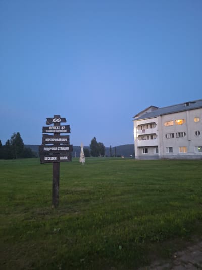 The image depicts an outdoor scene at dusk, likely in the city of Atamanovka, Russia. In the foreground, a wooden signpost with multiple directional arrows stands on a grassy field. The arrows indicate "ПРОКАТ #2 ТЕХНИКИ И СНАРЯЖЕНИЯ" (Rental #2 Equipment and Gear), "БЕРЕГОВОЙ ПАРК" (Coastal Park), "ЛОДОЧНАЯ СТАНЦИЯ" (Boat Station), and "БЕСЕДКИ ПЛЯЖ" (Gazebos Beach). To the right of the signpost, a white umbrella with a striped pattern is partially visible, suggesting an area for relaxation or recreation.
In the mid-ground, a multi-story building with a grey roof and light-colored facade is visible. Several windows on the building are illuminated, indicating occupancy. The building appears to be an apartment complex or a similar residential/communal structure. The surrounding landscape consists of a well-maintained grassy area, with trees and hills visible in the background under a clear, pale blue sky. The overall impression is of a quiet, possibly recreational, area in a rural or semi-rural setting, as twilight descends.