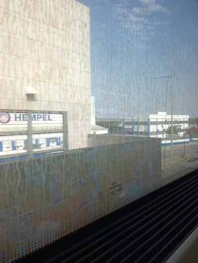 The image captures a view from inside a vehicle, likely a train, looking out at a modern industrial or office complex. The foreground is dominated by the interior of the vehicle, with dark, ribbed blinds running horizontally across the bottom. The window, which shows signs of dirt and smudging, offers a distorted view of the exterior.

On the left, a large, light-colored building with horizontal paneling is visible. A prominent sign with a red and blue circular logo and the word "HEMPEL" in blue lettering is mounted on the facade of a building within this complex. This sign is framed by horizontal metal bars, suggesting it's visible through a fenced or grated area. Behind this, another building with blue and white accents and rows of windows can be seen.

Further in the distance, a long, white, multi-story building with a blue stripe running along its roofline and blue window frames is situated. A single street lamp with a modern design stands to the right of this building against a bright, slightly cloudy blue sky. Trees with green foliage are scattered around the buildings.

The environment appears to be an industrial or commercial area, likely part of a business park or industrial zone. The clear sky suggests fair weather. The overall impression is one of a functional, modern infrastructure.

Based on the presence of the "HEMPEL" signage, it is highly probable that this scene is located in the vicinity of the Hempel factory or distribution center in Palmela, Portugal.