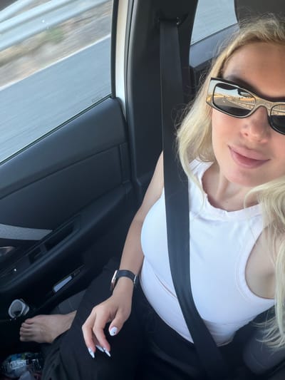 A woman with blonde hair and sunglasses is in the passenger seat of a car, traveling on a highway. The car's interior is visible, including the door panel, seatbelt, and part of the dashboard. The woman is wearing a white tank top and black pants. Her left foot is visible, bare and resting on the floor. Her right hand is on her lap, showing white-tipped fingernails. The reflection in her sunglasses shows the highway and a clear sky. The scene is consistent with travel on a highway, and the image appears to have been taken during daylight hours. The location of Villarrubio, Spain, is a plausible setting for such a scene, given the road infrastructure and general environment depicted.
