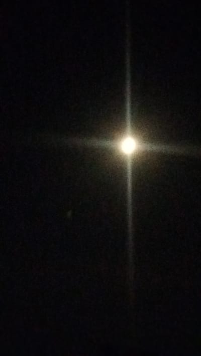 A bright, circular light source, likely the moon, is visible in the dark night sky. It is surrounded by a bright halo and a vertical streak of light extends above and below it, suggesting it is a primary light source. There are some faint, out-of-focus specks in the background. The image is grainy and dark, with no discernible features of the city of Kostiantynivka, Ukraine.