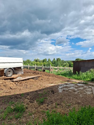 The image depicts an outdoor farm scene, likely in Косяково, Russia, under a cloudy sky with patches of blue. In the foreground, a large pile of brown material, possibly manure or soil, is visible, interspersed with dry straw and some green grass. To the left, the rear of a white trailer is parked on wooden planks, with a tire and part of its undercarriage visible. Beyond the pile of material, a wooden fence runs horizontally across the frame, with a gate-like structure in the middle. Behind the fence, a dense line of green trees forms the horizon, with a dark brown wooden shed or structure to the right. The ground is a mixture of dirt, straw, and vegetation, suggesting an agricultural or animal husbandry setting.