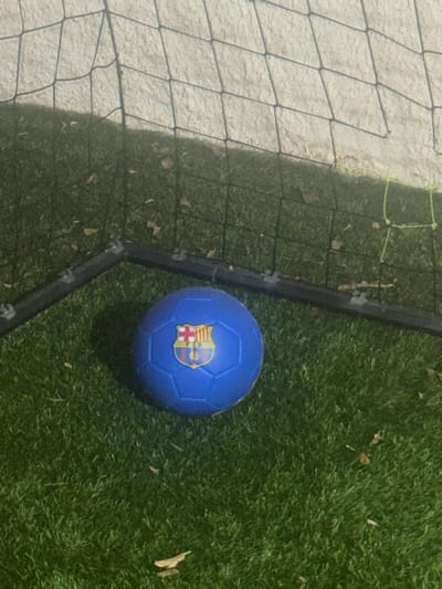 A blue soccer ball with the FC Barcelona logo is resting on artificial turf near a net. The scene appears to be a soccer goal area on a grassy field, likely in a park or sports facility. The background shows a textured surface and a portion of a net, suggesting an outdoor setting. The lighting indicates it might be evening or a dimly lit area. There is no indication of specific location in Hadera, Israel.
