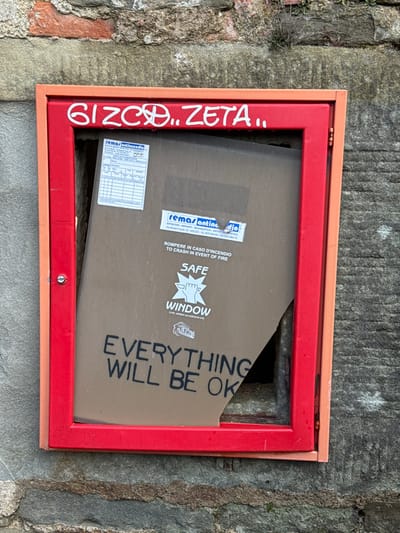 A broken fire emergency box is depicted mounted on a weathered stone wall in the city of Cortona, Italy. The rectangular box features a prominent red outer frame and a peach-colored inner frame. The front transparent panel, appearing dirty and tinted brown, has a significant triangular section missing from its bottom right corner, revealing the dark interior. Printed on the remaining panel are multiple white labels, including "remas antincendio" branding and what resembles an inspection chart. Below these are instructions in black text: "ROMPERE IN CASO D'INCENDIO" (Break in case of fire) and "TO CRASH IN EVENT OF FIRE." Further down, a white graphic shows a stylized fist breaking a window, labeled "SAFE WINDOW," with "TYPE APPROVAL CERTIFICATE" below it. At the bottom of the panel, large black letters spell out "EVERYTHING WILL BE OK." White graffiti, reading "GIZCO...ZETA..", is visible on the top section of the red frame. The irregular stone wall backdrop shows grey and brown tones with some mortar lines and sparse green vegetation at the top. No individuals are present in the scene.
