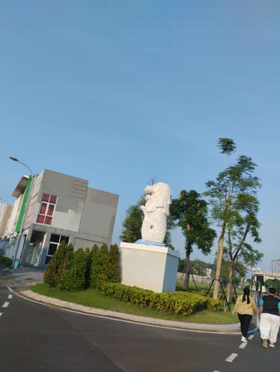In the city of Sidodadi, Indonesia, a Merlion statue stands proudly on a white pedestal with a blue wave design at its base. The statue, a mythical creature that is half lion and half fish, is a symbol of Singapore but has been replicated in various locations, including this one in Sidodadi. Beside the statue, lush green trees and manicured bushes surround the area, adding to the serene ambiance.
In the background, a modern, grey building with red-framed windows stands tall, its unusual tilt adding a unique architectural element to the scene. The building's structure suggests it might be an office or commercial space, with a green trim along its side and a lamppost positioned in front.
Two individuals, a man and a woman, are captured walking away from the camera on a paved road. The woman, dressed in a light brown jacket and black pants, has her hair covered by a black hijab. The man is seen in a black vest over a light-colored shirt and white pants. They appear to be enjoying a leisurely stroll, possibly for exercise, against the backdrop of the clear blue sky. The road they are walking on is marked with white dashed lines, indicating it's a pathway for pedestrians or a light traffic area.