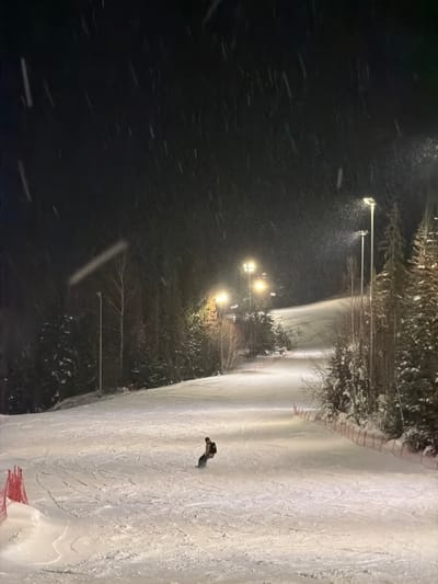 This image depicts a nighttime scene at a snow-covered ski slope in Makarovo, Russia, during active snowfall. A single individual, dressed in dark clothing and wearing a backpack, is visible snowboarding down the groomed slope, appearing to be in the process of turning left. The slope is illuminated by a series of at least five tall, bright floodlights positioned along the right side, casting a yellowish-white light on the snow. Snow-covered evergreen and bare deciduous trees line both sides of the slope, bordering the forested area. Orange safety netting is present along the lower right edge of the slope. Numerous snowflakes are visible in the air, captured by the artificial illumination. The overall environment is a wintery, mountainous or hilly landscape, operating as a lit ski facility at night.