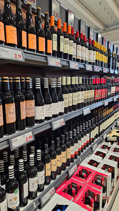 The image depicts a retail display of wine bottles on shelves in a store. The bottles are primarily red wine and are arranged in rows. Some bottles have prominent labels with prices displayed below them. The shelves are made of metal and are gray. The background consists of a white ceiling with visible lighting fixtures and possibly other retail displays. The overall environment suggests a supermarket or liquor store. There are no people visible in the image.
The specific location in Cuarte de Huerva, Spain, is an aspect that cannot be determined from the visual information alone. The provided image contains no explicit or implicit clues that would allow for its localization to that particular city. Therefore, the description is limited to the observable elements within the image.