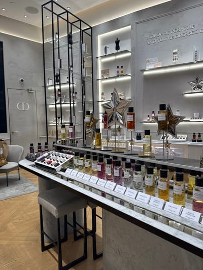 The image displays an unoccupied interior of a Dior fragrance boutique, located in Saint-Laurent-du-Var, France. The primary subject is a long, curved counter with a white marble top, dominating the foreground and midground. This counter showcases multiple rows of perfume bottles, each identified by a white label with its respective name, such as "GRIS DIOR," "AMBRE NUIT," and "ROUGE TRAFALGAR." On the far left end of the counter, a mirrored tray displays an array of small cosmetic products, possibly lip glosses or polishes. Two grey upholstered bar stools with dark metal frames are positioned beneath the counter on the left. The flooring consists of light-toned wooden parquet.
In the midground, directly behind the main counter, several large, mirrored star-shaped displays hold individual perfume bottles on stands. To the left, a tall, black-framed shelving unit with clear glass tiers exhibits more perfume bottles and small black torso mannequins. Further right, against the light grey textured wall, are wall-mounted shelves with integrated lighting, also displaying various fragrance products and decorative items. An embossed inscription on the rear wall reads: "A DROP OF PERFUME AND YOU'RE DRESSED IT." On the far left of the scene, a light-colored door featuring the "CD" logo is visible, with a white security camera mounted above it. In the far left background, a grey upholstered armchair and a brown paper bag are present. The environment is brightly illuminated by recessed ceiling lights and integrated shelf lighting, creating a clean and luxurious retail aesthetic. No individuals are present in the scene.