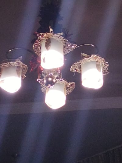 The media file shows a chandelier with four lights hanging from a ceiling. The chandelier has a dark metal frame with decorative elements. Each light is enclosed in a cylindrical lampshade with an ornate, patterned trim around the top. The lights are on and emit a bright, warm glow, creating light beams that streak through the dimly lit space. The background is dark and appears to be a ceiling with some decorative elements visible, possibly a dark textured surface. There is a hint of a dark, possibly wooden surface below the chandelier. The lighting suggests it could be evening or indoors with the lights on for illumination. The scene is an interior shot, likely in a room or hallway, given the presence of the chandelier and the ceiling detail. No people are visible, and the focus is solely on the lighting fixture. There is no visible text in the image.