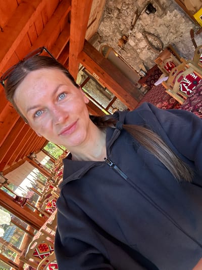 In Gusinje, Montenegro, a woman with blue eyes and a braid is taking a selfie in what appears to be an indoor seating area with exposed wooden beams and stone walls. She is wearing a dark zip-up hoodie. The background shows rows of chairs with red and white patterned upholstery, suggestive of a traditional restaurant or guesthouse. Sunlight streams in from windows, illuminating the space and casting shadows.