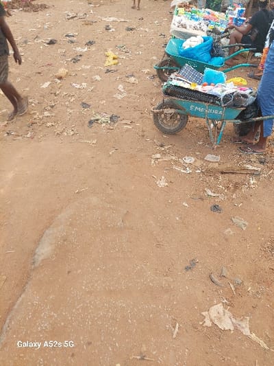 The image depicts an outdoor market scene in Awkuzu, Nigeria. The ground consists of unpaved, reddish-brown dirt, extensively scattered with various debris, including black, yellow, and white plastic bags, as well as miscellaneous paper and refuse.
On the left side of the frame, a person is partially visible from the lower thighs down, wearing dark shorts and flip-flops, in the act of walking from left to right across the dirt ground.
On the right side, two teal-colored wheelbarrows are positioned, functioning as vendor stalls. The front wheelbarrow bears the inscription "EMENUGA" in white lettering on its side and is laden with various merchandise, including patterned textiles, light blue fabric, and spherical reddish produce. The second wheelbarrow, situated behind the first, contains a blue plastic bag filled with white items and other packaged goods.
Behind these wheelbarrows, two individuals are partially visible, seated. One individual, wearing dark clothing and flip-flops, is positioned near the front wheelbarrow. A second individual, also in dark clothing, is seated further back amidst a larger display of packaged commodities and produce arranged on a raised surface or table. The overall scene is illuminated by natural daylight. The watermark "Galaxy A52s 5G" is visible in the lower-left corner of the image.