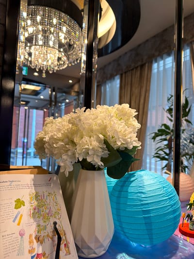 The image depicts an indoor scene with several decorative elements. In the foreground, a white vase holds a large bouquet of white hydrangeas. To the right of the vase, a bright turquoise paper lantern rests on a blue, shimmering surface. Behind the hydrangeas and to the left, a book with illustrations is open, showing text and images related to "Tanabata Decorations." The reflections in the mirrored background reveal a large, ornate crystal chandelier and the interior of what appears to be a room with draped curtains and other decorative items. The book's content suggests a connection to Japanese culture, specifically the Tanabata festival. The scene is likely set in a hotel, restaurant, or retail space in Chuo, Japan, given the cultural context and the presence of such decorations. No people are visible in the image.