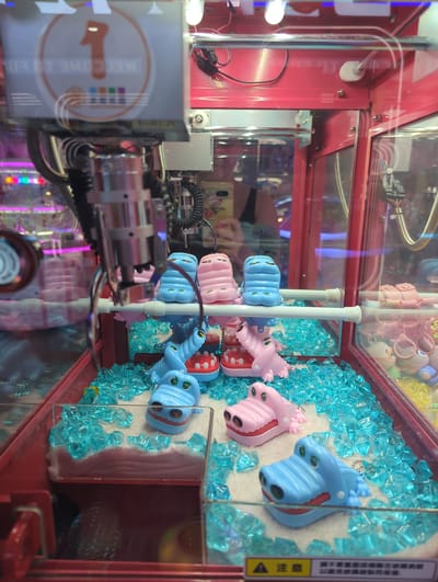 The image shows the interior of a claw machine toy dispenser located in Xishan District, China. The machine is bright pink and filled with various small toys. In the foreground, several blue and pink toy alligators are scattered among translucent blue decorative "ice" pieces, some of which are on a fluffy white surface. A metal claw mechanism is visible hanging from above, positioned over the toys. In the background, a screen displays the number "1" inside a circle and the text "WELCOME TO FUN". Other colorful toys and lights are visible through the glass of the machine. A person's reflection is partially visible in the glass.
