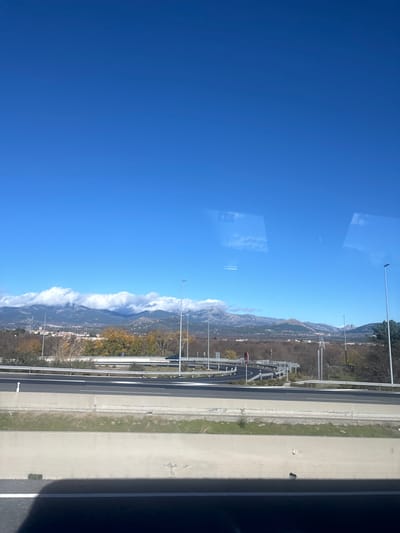 This landscape photograph captures a vast outdoor scene featuring a highway, mountains, and a clear blue sky. The image appears to have been taken from inside a vehicle, as indicated by faint rectangular reflections and distorted clouds visible on a window pane, particularly in the upper sky area.

In the foreground and lower section of the image, a multi-lane highway extends horizontally across the frame. Its grey surface is marked with white lines, and it is bordered by guardrails and tall, white streetlights. A strip of green, grassy land with scattered trees and shrubs separates the highway from the more distant elements.

The mid-ground is dominated by a prominent mountain range that stretches across the image. These rugged mountains are covered in varied vegetation, and their peaks are partially shrouded in low-lying white clouds or mist, suggesting either high altitude or specific weather conditions.

The upper portion of the photograph is filled with a vibrant, clear blue sky, indicating bright and sunny weather. The time of day is clearly daytime, likely morning or early afternoon, given the strong illumination. No specific human activity, individuals, or legible text are visible. The scene conveys a sense of expansive natural beauty coexisting with modern infrastructure, characteristic of the mountainous region around Guadarrama, Spain.