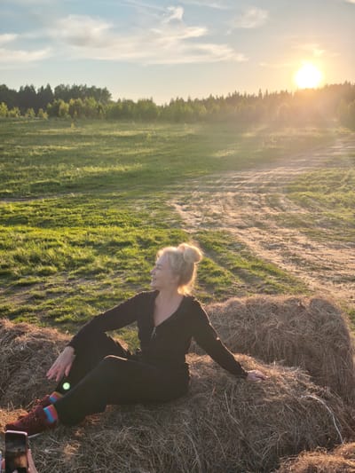 This vertical image captures a serene outdoor scene during the golden hour, likely in Стеклянный, Russia, as the sun sets. The warm, golden light of the low sun emanates from the upper right, casting a strong lens flare and illuminating the landscape.

The main subject is a woman, appearing middle-aged, with blonde hair styled in a messy bun. She is sitting comfortably on a large, round bale of hay, positioned towards the lower center of the frame. She wears a dark, possibly black or dark brown, long-sleeved jumpsuit or tracksuit, accented by vibrant, multi-colored striped socks and dark sneakers. Her head is turned to the left, and she has a gentle, contented smile on her face, suggesting a relaxed and happy mood.

The setting is an open, rural field, characterized by patches of green grass and bare earth or dirt paths. A dense line of trees forming a forest marks the horizon in the background, under a partly cloudy sky. The low angle of the sun creates long shadows and highlights the textures of the hay bale and the field.

In the bottom left corner, a smartphone held by another individual is partially visible, indicating that the woman is posing for a photograph. The overall atmosphere is peaceful and natural, showcasing a tranquil moment at sunset. No visible text is present in the image.