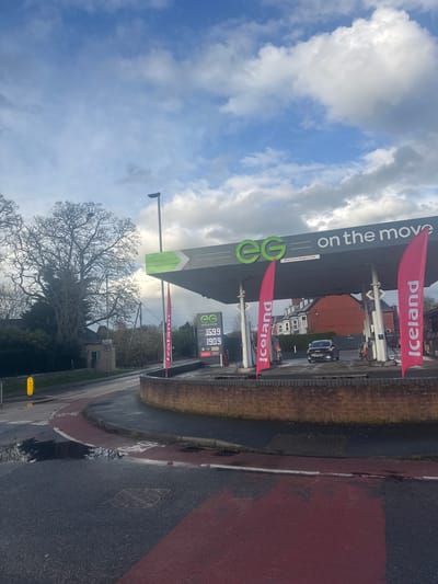 This outdoor shot is taken in Welshpool, United Kingdom, focusing on an EG fuel station with Iceland food market affiliation. The fuel price display shows gasoline at 159.9 and diesel at 190.9. "Iceland" flags flank the EG-branded fuel station canopy. The sky is partly cloudy. Street furniture, including a lamppost and traffic control equipment, are present. A brick wall borders a red-marked pavement and road surface. A large tree with bare branches is positioned in the foreground.