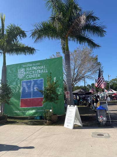 An outdoor scene captures a sunny day at what appears to be the US National Pickleball Center, likely during an event like the US Open Pickleball Championship. The image is taken from a low angle, looking up, with prominent palm trees framing the left side.
A large green banner dominates the center of the frame, displaying text that reads, "WELCOME TO THE... US NATIONAL PICKLEBALL CENTER" with a logo featuring "US P" and a pickleball icon. Below this, partially obscured, is "HOME OF THE US OPEN PICKLEBALL CHAMPIONS". Small trees are planted in front of the banner.
To the right of the palm trees and behind the banner, a lively event area is visible. Several American flags are displayed on poles, flapping in the breeze. Various white and black tents or stalls are set up, with one black tent clearly showing the brand "ROKNE". People are seen milling around these tents, suggesting vendors, information booths, or spectator areas.
A red "NO PARKING" sign with an arrow pointing left is attached to a pole. A white A-frame sign stands on a concrete path, displaying information, though much of the text is small. Legible snippets from OCR include "RECYCLING BIN", "BOTTLES AND CANS ONLY", and "WATER". A clear recycling bin filled with plastic bottles and aluminum cans is also visible near the path.
The setting is bright and sunny, indicated by the clear blue sky and distinct shadows cast on the concrete path. The presence of palm trees and the clear weather suggest a warm climate. The overall impression is that of a bustling, well-organized outdoor sporting event or festival.