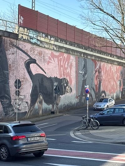 The image depicts an urban street scene at Adalbertsteinweg 28, Aachen, Germany, beneath a railway bridge. A long, brick retaining wall, extensively covered with a mural and graffiti, forms the left boundary of the road. The mural prominently features a large, dark-colored dog, possibly a Rottweiler or similar breed, on a leash, depicted in profile walking right. A partial human leg is visible to the dog's left. Large, red-outlined graffiti tags, including "THEK," are present on the mural. Above the brick, a gray concrete band precedes a red-brown slatted sound barrier fence, with railway tracks and overhead power lines visible against a clear blue sky.
The asphalt road below features white lane markings and a red-painted section, suggesting a bicycle lane or dedicated area. An intersection or road curve is visible. A dark gray Audi A3 Sportback, bearing the German license plate "DN UD 99," is positioned in the foreground, facing away from the viewer. Further along the road, a dark blue hatchback and a silver Mercedes-Benz coupe are visible. A dark gray bicycle is parked upright against a silver pole with a red-and-blue circular "No Stopping" sign. Another black pole with two circular signs, including a "No Stopping" variant, stands on the left near the mural. Bare tree branches are visible against the clear sky. The scene is illuminated by natural daylight.