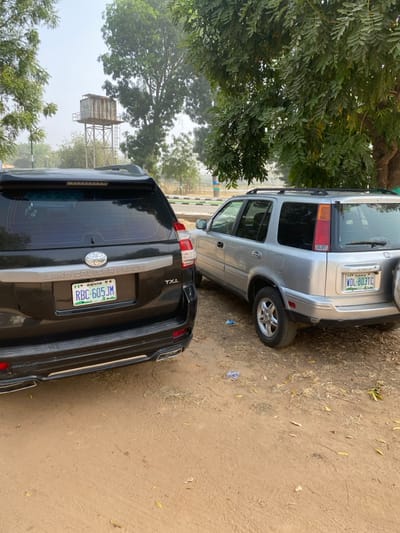 The scene in Yan Dala, Nigeria, features two sport utility vehicles parked on a dry, reddish-brown dirt surface. On the left, a black Toyota Land Cruiser Prado TXL is positioned with its rear facing the viewer. It displays an Abuja-registered Nigerian license plate: "RBC-605 JM." The vehicle has a tinted rear window, roof rails, and chrome-tipped dual exhaust outlets. To its right, a silver Honda CR-V, likely a first-generation model, is visible from its rear passenger side. This vehicle exhibits a Kano-registered Nigerian license plate: "WDL-803TC," a rear-mounted spare tire, roof rails, and five-spoke alloy wheels.

The ground is predominantly bare earth with sparse patches of dried vegetation and scattered dry leaves. A crumpled blue plastic item is visible on the dirt near the front wheel of the silver CR-V. In the middle ground, behind the vehicles, a paved road with a green painted curb is present. The background is dominated by large, leafy trees under a hazy, bright sky. A tall, rusted metal water tower with a large, square tank is visible in the distance, positioned behind the black SUV and partially obscured by tree foliage. No individuals are present, and no actions or interactions are depicted.