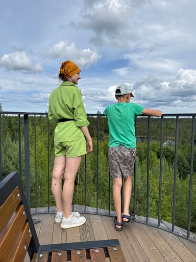 Two people, a young female and a young male, stand on a wooden platform, looking out at a forest landscape under a cloudy sky. The female, on the left, faces away from the camera and wears a green linen outfit consisting of a collared shirt and shorts, an orange bandana on her head, and white sneakers. The male, on the right, also faces away from the camera, wearing a green t-shirt, patterned shorts, a light-colored baseball cap, and dark sandals. They are positioned behind a black metal railing. In the foreground, a wooden bench with black metal supports is visible. The background is a dense forest with various shades of green trees and a body of water in the distance. The sky is filled with large, white cumulus clouds. The location is identified as the city of Peski, Russia.