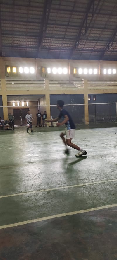 An indoor badminton court in Kutal, Sukon, Indonesia is depicted. A male badminton player in a dark blue t-shirt and white shorts is in motion on the court, in the process of striking a shuttlecock. His body is blurred due to the action. Another player, wearing a white and grey patterned t-shirt and black shorts, is also on the court, appearing to be observing or anticipating the shuttlecock's trajectory. Several other individuals, likely spectators or fellow players, are visible in the background, standing or sitting near the perimeter of the court. The court itself has a green floor with white lines marking its boundaries. The structure features a high, tiled roof supported by beams and illuminated by rows of square windows that allow light to enter the space.