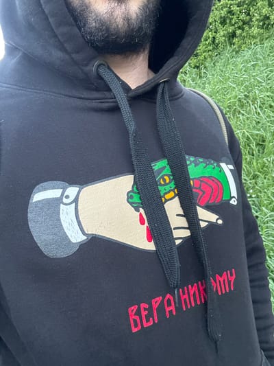 A man wearing a black hoodie lies on grass. The hoodie has a graphic of a hand holding a grenade. Vertical red text in Cyrillic script is next to the graphic. A black hood is visible on the left side of the frame. The man's face is obscured by the hood, revealing only his beard. The grass is green and appears to be well-maintained. The location cannot be definitively determined as being in Welshpool, United Kingdom based on the visual information provided.