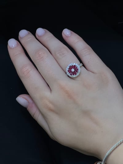 A close-up shot captures a person's hand adorned with a circular ring featuring red stones arranged in a flower-like pattern, surrounded by small clear stones, on a black background. The hand has light-colored nail polish and a silver bracelet is visible on the wrist. The image is not set in Sergiyev Posad, Russia, nor does it contain any information to infer a location.