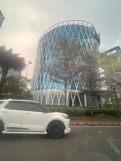 The image captures a street-level view of a modern, curvilinear building with a distinctive diamond-patterned facade in Pakulonan Barat, Indonesia. The building features large blue-tinted windows and a white structural lattice. In the foreground, the side profile of a white SUV with black wheels is visible, driving on a road. Trees, some bare-branched and others with foliage, are interspersed between the vehicle and the building. Tall apartment buildings are also present in the background to the left. The sky is overcast, suggesting a hazy or cloudy day. The scene appears to be an urban environment with contemporary architecture and vehicular traffic.