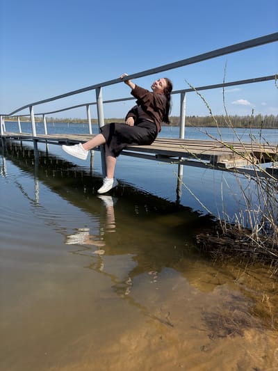 A girl in a brown jacket and black culottes is hanging off the railing of a wooden pier extending into a body of water. She is wearing white sneakers. The pier has metal railings. Her reflection is visible in the water. The water is murky and reflects the pier and surrounding vegetation. The sky is blue. Crokhovo, Russia is not discernible from the image.