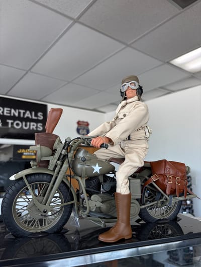 A realistic action figure of a man in vintage military attire, complete with a helmet, goggles, and a tan uniform, is posed riding a detailed replica of a vintage olive-drab motorcycle. The motorcycle is equipped with leather saddlebags and a rifle case mounted on the front. The action figure and motorcycle are displayed on a reflective black surface, possibly a display case, in what appears to be an indoor commercial space. In the background, signage can be seen, including a black sign with white text reading "RENTALS & TOURS" and a smaller yellow sign with "JACKE RENTAL." A Route 66 sign is also visible. The ceiling features suspended acoustic tiles and fluorescent lighting, typical of a retail or office environment. The scene is illuminated by overhead lighting, casting a bright light on the subjects and creating reflections. The overall impression is a curated display of a historical military scene. The location is identified as Des Plaines, United States of America.