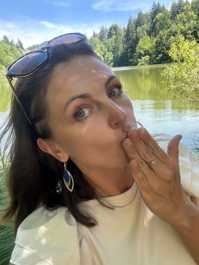 A woman is blowing a kiss to the camera with her right hand cupped over her mouth. She is wearing a light-colored shirt, dangling earrings, sunglasses on her head, and a ring on her right ring finger. She is in an outdoor setting with a body of water and trees in the background, likely near Ravilloles, France.