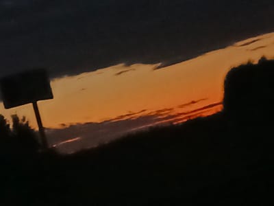 The image shows a silhouette of a signpost against a dark sky with an orange and yellow sunset. The signpost appears to be a dark, rectangular shape supported by a pole, with a slightly blurry and textured surface. The sky is dominated by the vibrant hues of the sunset, with clouds creating streaks of light orange and yellow against the darker parts of the sky. The lower portion of the image is completely black, representing the silhouette of the landscape or ground. The overall impression is of a rural or roadside scene during dusk. There is no indication of the specific location within Shestunikha, Russia, beyond the fact that it is a sunset scene.