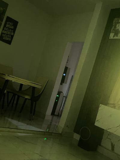 This image captures an interior scene, likely a residential or commercial building, located in Okpanam, Nigeria. The environment is dimly lit with a pervasive green hue. In the left foreground, a dining area is visible, featuring a table with a light-colored surface and dark, thin legs accented with gold. Two matching chairs are positioned around it. Above the dining area on the wall, a rectangular black framed poster displays white text: "STAY STRONG BELIEVE IN YOURSELF NEVER GIVE UP."
Towards the center and right, an open doorway or hallway leads deeper into the structure. Within this passage, two vertical electronic devices are mounted, each displaying active green indicator lights. The upper device also shows blue indicator lights. The floor consists of light-colored, possibly polished, tiles which reflect the green lights from the devices. A prominent white column or architectural feature separates the main room from the hallway on the right. Below this column, a dark, rectangular object with a circular feature (possibly a speaker) is partially visible. The wall above this object is adorned with white, marble-patterned paneling or tiles. No individuals or specific actions are depicted.