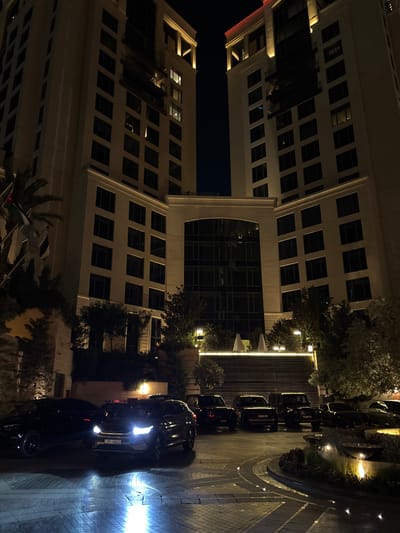 The image captures a nighttime view of two tall, modern buildings in Amman, Jordan. The buildings, illuminated by exterior lighting, appear to be a hotel or luxury residential complex. The foreground is dominated by a row of parked black vehicles, including sedans and SUVs, with their headlights on. The vehicles are positioned on a patterned paved surface that reflects the light from the cars and surrounding lamps. In the background, there are trees and some outdoor seating areas with umbrellas, also lit by subtle lighting. Flags are visible to the left of the buildings, suggesting an international or official presence. The overall scene conveys a sense of luxury and nighttime activity in an urban environment.