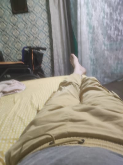 A person is lying on a bed, with their legs extended towards the camera. The person is wearing tan pants and a grey t-shirt. The bed has a yellow and white checkered blanket. In the background, there is a wall with a blue and white patterned wallpaper, and a white curtain. To the left of the curtain, there is a dark object that appears to be a piece of furniture or a bag. The image is taken from a low angle, looking up at the person's legs. The overall scene suggests a casual or relaxed indoor setting. The city of Saransk, Russia, is not visible or suggested in this image.