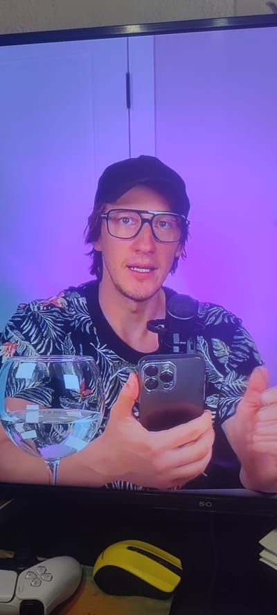 The media file shows a man wearing a black baseball cap and glasses. He is dressed in a black t-shirt with a tropical leaf pattern. The man is holding a smartphone in his right hand and appears to be speaking into a microphone attached to the phone. A glass of water is visible to his left. The background is illuminated with a purple and blue gradient light. In the foreground, a white video game controller and a yellow computer mouse are placed on a dark surface. The setting appears to be indoors, possibly a home studio or office, given the presence of recording equipment and gaming peripherals. The lighting suggests it could be evening or a dimly lit indoor space. The man's expression is neutral, and he seems engaged in a recording session or a live stream. The visible text on the screen edge is "SO", likely a brand logo. The overall atmosphere is casual and focused on content creation.